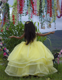 Young girl in a yellow dress standing in front of hanging floral decorations with a scenic background. by kids fashion designer in pune