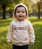 Child wearing a beige hoodie with 'MLMNZY' text in a park setting  by fashion designers in pune